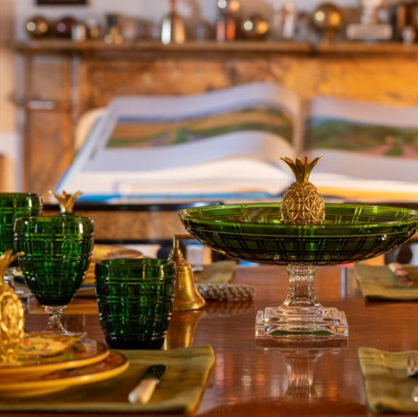 A styled dining table setting with multiple green glass goblets, gold and white plates, a large green pedestal bowl with a pineapple topper, and matching green and blue pineapple accents, photographed in a warm indoor room with a blurred book and lamp in the background.